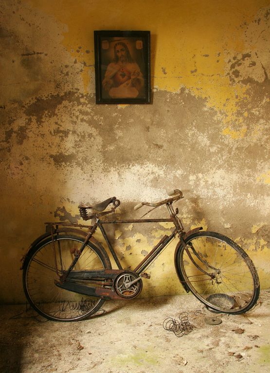 An old bike propped up against a wall under a picture of the Sacred Heart. The bicycle still has its pump attached.