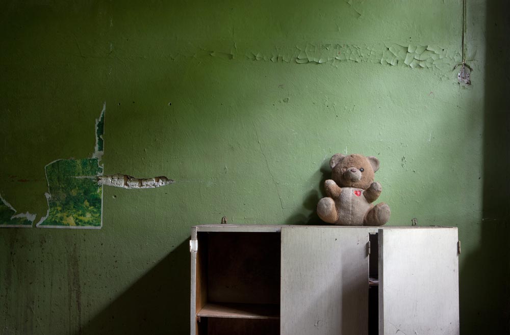 In the bedroom of an abandoned home a forgotten Teddy Bear sits on top of a dressing table